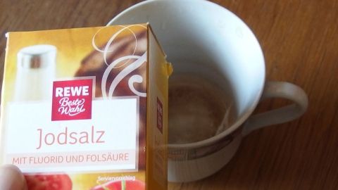 Eine Packung Jodsalz wird über eine leere Tasse gehalten, sichtbar auf einer Holzoberfläche, als ob jemand sie benutzen möchte.