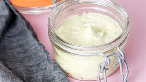 Ein Glas mit selbstgemachter Butter steht auf einem rosa Hintergrund neben einem grauen Geschirrtuch.