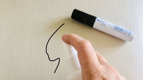 Eine Hand sprüht Reinigungsmittel auf eine von Edding beschmutzte Polsterfläche, während ein Marker daneben liegt.