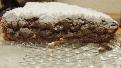 Ein Stück Mandelkuchen liegt auf einer dekorativen Platte und wird von Puderzucker bestäubt, umgeben von einer rustikalen Kücheneinrichtung.