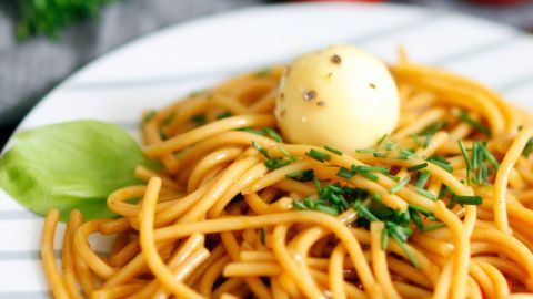 Ein Teller Spaghetti mit Trüffelbutter, dekoriert mit einem Butterball und frischen Kräutern, auf einem Tisch mit roten Tomaten.