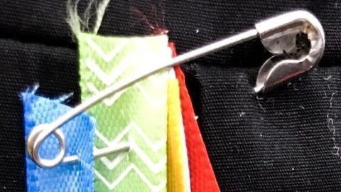 Bunte Reste von Geschenkband sind auf einer Sicherheitsnadel aufgefädelt, vor schwarzem Hintergrund angespannt.