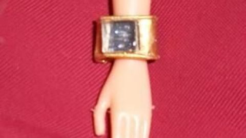 Armband für die Barbie aus Tütenverschluss hergestellt Ein goldenes Armband aus einem Tütenverschluss schmückt das Handgelenk einer Barbie-Puppe auf rotem Untergrund.