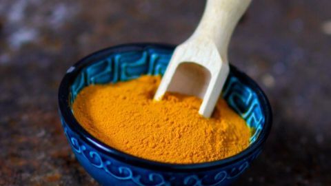 Eine kleine blaue Schüssel enthält leuchtend gelbes Kurkuma-Pulver, während ein Holzlöffel darüber hängt.