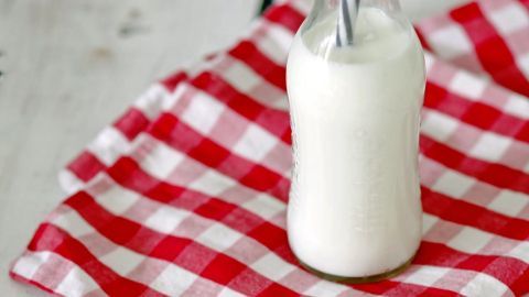 Eine Flasche Mandelmilch mit einem Strohhalm steht auf einem karierten Tuch, umgeben von Mandeln auf einem Holztisch.