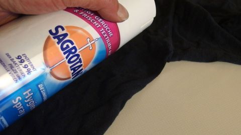 Eine Hand hält ein Desinfektionsmittel über einem schwarzen Kleidungsstück, um Schweißgeruch zu entfernen.