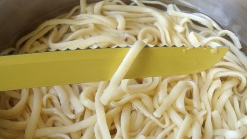 Mit einem gezackten Messer einzelne Spaghetti zur Garprobe aus dem Wasser heben. Ein einzelnes Spaghetti wird mit einem gelben Messer aus einem Topf mit kochendem Wasser gehoben, um die Garprobe zu testen.