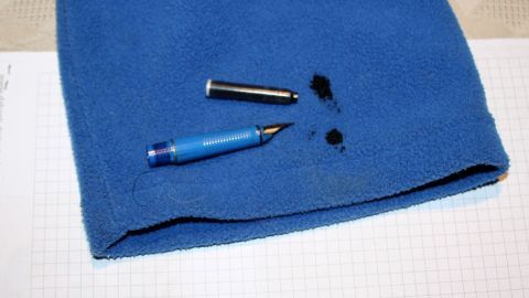 Wenn man Tintenflecken in der Kleidung hat, kann man sie einfach mit einem Tintenkiller entfernen - den Fleck einfach aufsaugen, das Kleidungsstück sofort abwaschen und weg ist er. Den Tintenkiller nicht zu lange einwirken lassen. Auf einem blauen Kleidungsstück liegen zwei Stifte und ein Tintenfleck, was das Problem des Tintenflecken Entfernens illustriert.