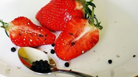 Saftige Erdbeeren liegen auf einem weißen Teller, teilweise aufgeschnitten, mit schwarzem Pfeffer daneben.