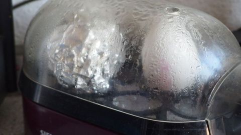 Kochendes Wasser in einem Eierkocher umgibt ein geplatztes Ei in Silberfolie und ein ganzes Ei, beschlagenes Glas.