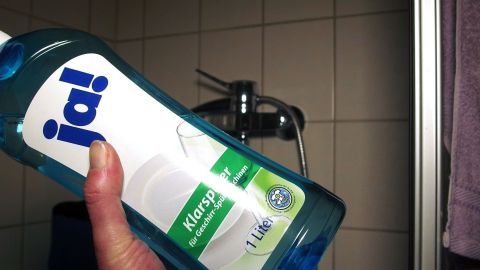 Eine Hand hält eine Flasche Geschirrklarspüler vor einer Duschwand, bereit zum Reinigen.