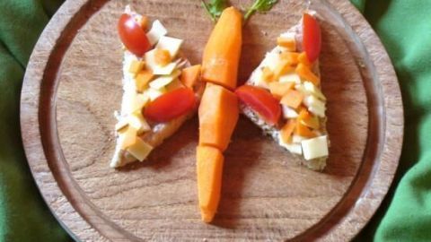 Käsetoast in Form eines Schmetterlings liegt auf einem Holzbrett, mit Tomaten und Möhre als dekorative Elemente.
