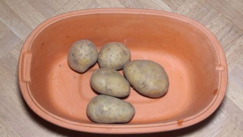 Sechs rohe Kartoffeln liegen in einem Römertopf, bereit zum Kochen ohne Wasser auf einer holzfarbenen Fläche.