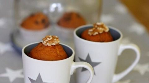 Zwei Tassen-Muffins stehen auf einem dezent gestalteten Tisch, während im Hintergrund eine Glasglocke weitere Muffins schützt.