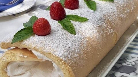 Eine Biskuitrolle liegt auf einem Teller, gefüllt mit Sahne und dekoriert mit Himbeeren und Minzblättern.