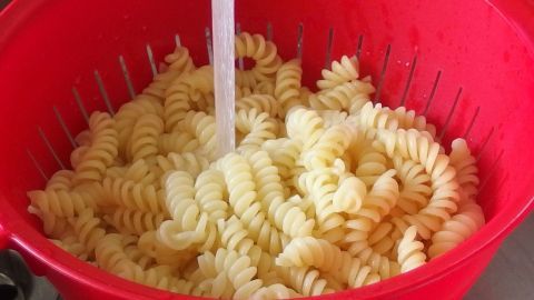 Pasta wird unter fließendem Wasser in einem roten Sieb abgewaschen, während sich in der Küche eine Spüle im Hintergrund befindet.