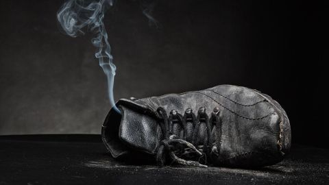 Ein abgenutzter Schuh liegt auf einem dunklen Untergrund, während Rauch aus der Öffnung aufsteigt, was stinkende Schuhe symbolisiert.
