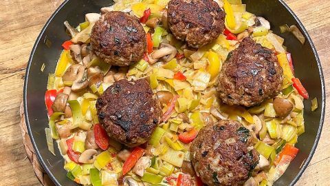 Kleine Frikadellen ruhen auf einem bunten Gemüsebett aus Lauch, Paprika und Pilzen in einer Pfanne auf Holz.