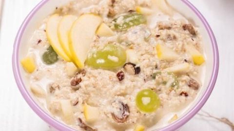 Eine Schüssel Original Schweizer Bircher Müsli mit Haferflocken, Trauben, Apfelstücken und Nüssen auf einem weißen Tisch.