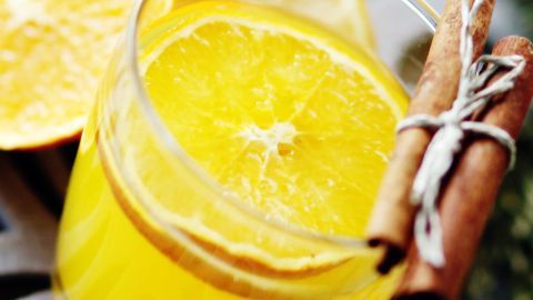 Ein gelbes Kinder-Cocktail-Glas mit einer Orangenscheibe und Zimtstangen, umgeben von frischen Zutaten und natürlichem Licht.