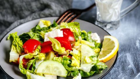 Ein frischer Salat mit verschiedenen Gemüsesorten liegt auf einem Teller, während ein Glas Joghurtdressing daneben steht.