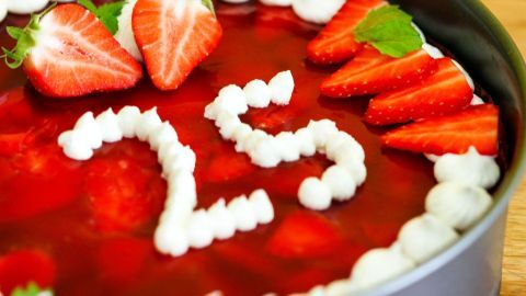 Ein rundes Obsttörtchen mit Erdbeeren und Tortenguss zeigt die Zahl "25" aus Sahne, auf einem Holzuntergrund.