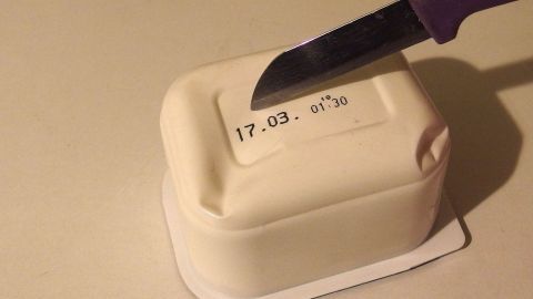 Eine Quark-Plastikschale liegt auf einem Tisch, während ein Messer die Folie oben ansticht, um den Quark herauszunehmen.