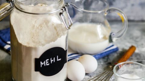 Ein Glas mit Mehl steht auf einer Arbeitsfläche, umgeben von Eiern, einer Schüssel Zucker und einer Glasflasche Milch.