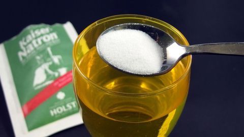 Ein Löffel Speisenatron schwebt über einem Glas mit gelber Flüssigkeit, während ein grüner Beutel im Hintergrund sichtbar ist.