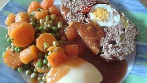 Gefüllter Hackbraten liegt auf einem Teller, umgeben von kürbis, erbsen und einer Kartoffel, in brauner Soße serviert.