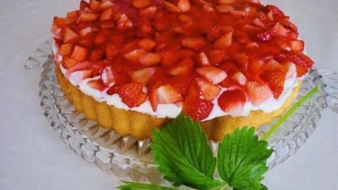 Eine Torte mit Erdbeeren und Tortenguss steht auf einem Glasplattenuntersetzer, umgeben von frischen Blättern.