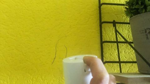 Den Strich mit Haarlack gut anspritzen, feuchtes Tuch darunterhalten um die blaue oder schwarze Tinte aufzufangen. Eine Hand hält eine Sprühdose, die auf eine gelbe Wand mit Kugelschreiberflecken zielt, während eine Pflanze im Hintergrund zu sehen ist.
