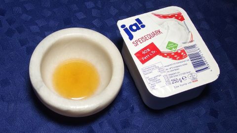 Eine kleine Schale mit Honig steht neben einem Quark-Becher, auf einem blauen Tisch bedeckt, bereit zur Gesichtsmaske.