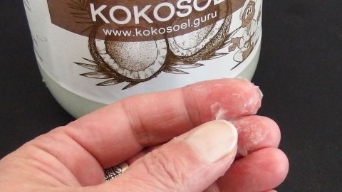Eine Hand hält eine kleine Menge Kokosöl, während im Hintergrund ein Glas mit der Beschriftung "Kokosöl" sichtbar ist.
