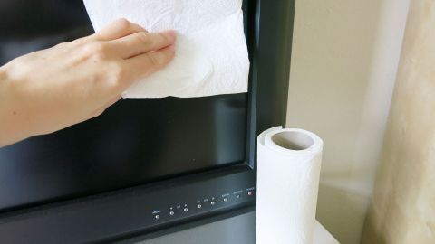 Ein Hand mit einem Papiertuch reinigt den Bildschirm eines Fernsehers, während eine Rolle Küchenpapier daneben steht.