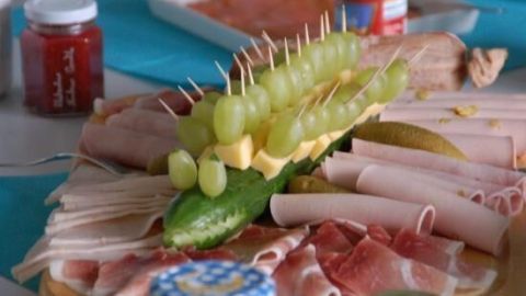 Ein Gurkenkrokodil mit Trauben und Schinkenstücken liegt auf einem kalten Buffet, umgeben von verschiedenen Leckereien.