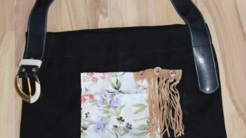 Eine Gürteltasche mit schwarzem Stoff, einer blumigen Tasche und einer Fransenverzierung liegt auf einem hellen Holzboden.