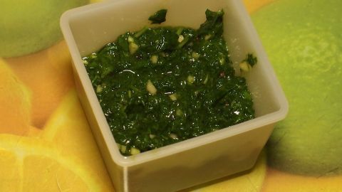 Ein kleiner, rechteckiger Behälter enthält grünes Pesto, das auf einem bunten Hintergrund mit Zitrusfrüchten steht.