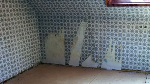 In einem Raum mit verblassten blauen Tapeten werden alte Tapeten von der Wand gelöst, während das Licht durch das Dachfenster fällt.