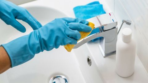 Eine Person reinigt einen Wasserhahn mit einem schwammähnlichen Tuch und Schmierseife in einem Badezimmer.
