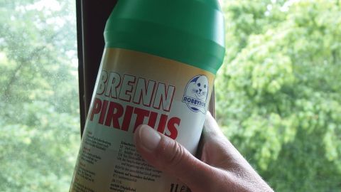 Eine Hand hält eine Flasche Brennspiritus, während sie ein Fenster mit Blick auf grüne Bäume reinigt, um Streifen zu vermeiden.