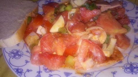 Ein bunter Tomatensalat mit Eiern und Bierschinken liegt auf einem Teller neben einer Scheibe Brot, umgeben von einer gemütlichen Umgebung.