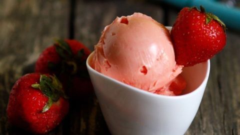 In einer kleinen weißen Schüssel liegt Erdbeer-Joghurt-Eis, umgeben von frischen Erdbeeren und einem Eislöffel.