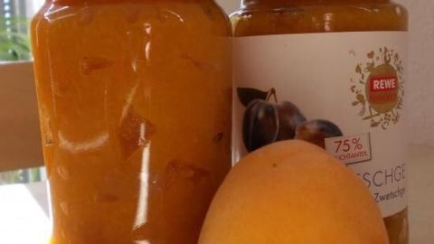 Gläser mit Aprikosenmarmelade stehen auf einem Tisch bei Sonnenlicht, ein Aprikose liegt davor. Text: REWE Feine Welt, 75% Fruchtanteil.