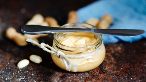 Ein Glas mit selbstgemachter Erdnussbutter auf braunem Untergrund, ein Messer darauf, umgeben von Erdnüssen.