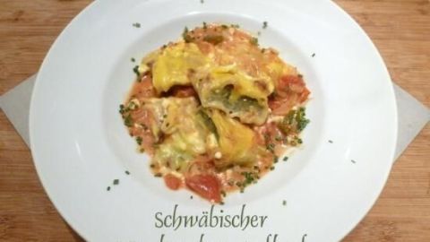 Maultaschen liegen auf einem Teller in cremiger Soße mit Tomaten und Kräutern, darunter steht „Schwäbischer Maultaschen-Auflauf“.