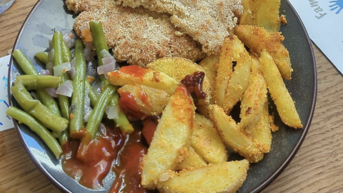 Ein Teller zeigt knusprige Haferflockenschnitzel neben grünen Bohnen und goldbraunen Kartoffelspalten, serviert auf einem Tisch.