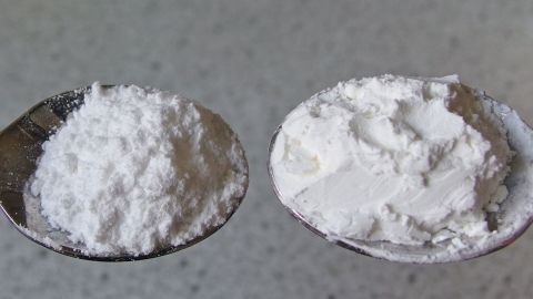 Ein Löffel mit pulverförmigem Sahnesteif steht neben einem Löffel mit körnigem Material auf einer blassen Oberfläche.