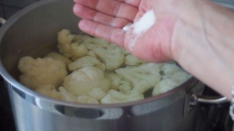 Blumenkohl wird in einem Topf mit Wasser gekocht, während Salz aus der Hand darüber gestreut wird, um die weiße Farbe zu erhalten.