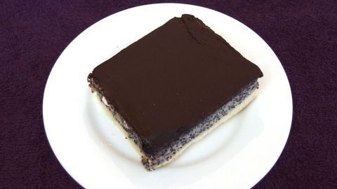 Ein Stück DDR Mohnkuchen liegt auf einem weißen Teller und zeigt eine dunkle, glänzende Schokoladenglasur, umgeben von einem lila Hintergrund.